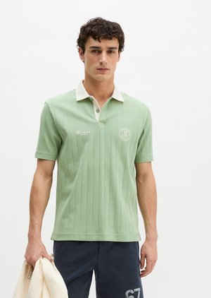 Homme portant un polo vert clair à manches courtes avec col blanc et pantalon bleu marine, tenant une veste blanche sur fond blanc.