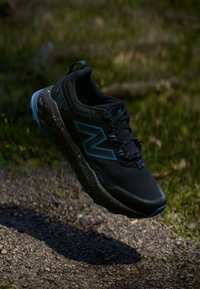 Chaussure de sport noire avec une tige texturée, des accents bleus, une semelle mouchetée et un système de laçage, conçue pour la durabilité et l'adhérence.