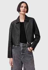 Chaqueta de cuero negra recortada con collar plano, cremallera frontal y mangas largas. Lleva puesta sobre una blusa negra y combinada con unos jeans grises de tiro alto.