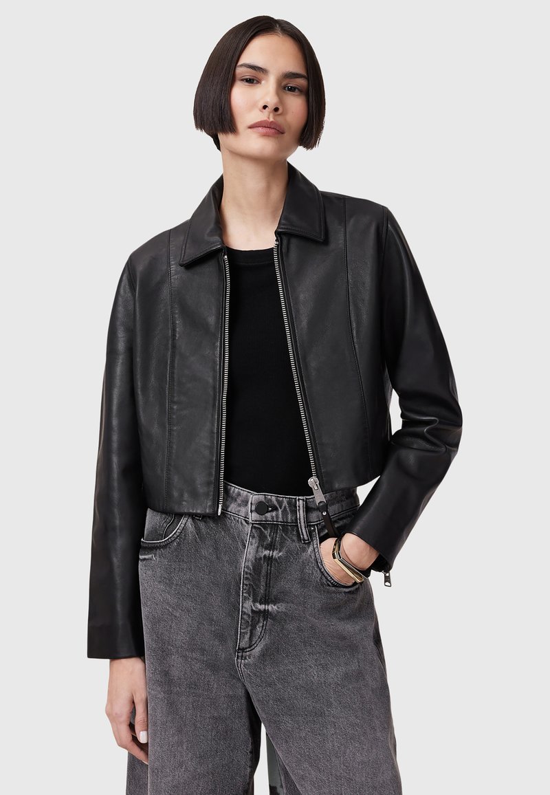 Chaqueta de cuero negra recortada con collar plano, cremallera frontal y mangas largas. Lleva puesta sobre una blusa negra y combinada con unos jeans grises de tiro alto.
