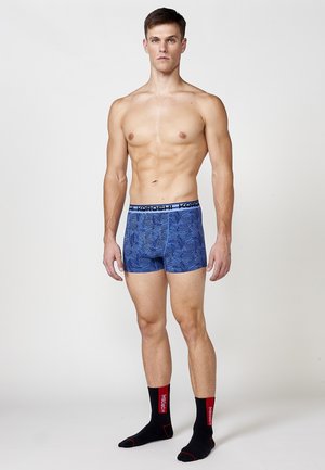 Blaue gemusterte Boxershorts mit einem elastischen Bund, die mit dem Branding "KOROSH" versehen sind, getragen mit schwarzen Socken mit roten Akzenten.