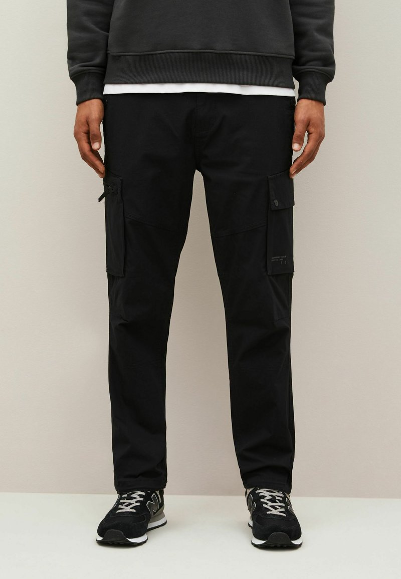 Svarta cargobyxor tillverkade av slitstark tyg, med flera sidofickor och en avslappnad passform. Matchade med svarta sneakers och en mörk sweatshirt.