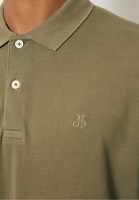 Detailní záběr na olivově zelenou bavlněnou polo košili s límcem, dvěma bílými knoflíky a malým vyšitým logem MOP na prsou.