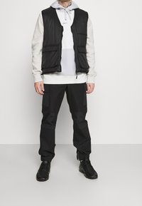 Svart vadderad väst över en grå hetsko hoodie, tillsammans med svarta cargobyxor. Funktioner inkluderar dragkedjor, fickor och en avslappnad design. Svarta sneakers fulländar looken.