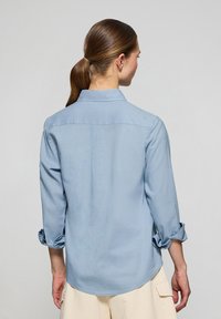 Camisa de botones azul claro con cuello y mangas de tres cuartos arremangadas. Tejido suave y ligero con un corte relajado y un discreto detalle de costura en la parte posterior.