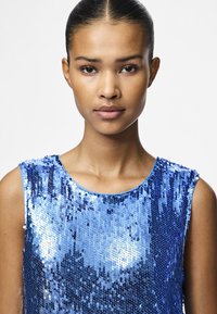 Top en sequins bleus avec un col rond et un design sans manches. Les sequins reflètent la lumière, créant une texture chatoyante sur le vêtement.