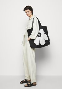 Black tote bag with a large white flower design, featuring sturdy fabric, wide straps, and a smooth texture. Model wears light-colored clothing.