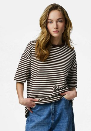 Fiatal nő hosszú, hullámos szőke hajjal, barna-fehér csíkos, laza ingben és kék farmernadrágban, kezét zsebében tartva áll.