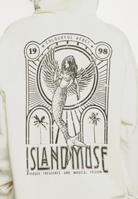 Sudadera con capucha color crema que presenta una gran estampa gráfica de una sirena con alas, flanqueada por palmeras y rodeada de texto y elementos decorativos.