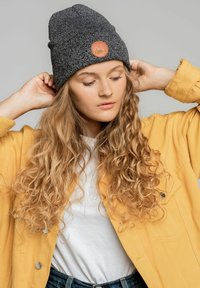 Czarna dzianinowa czapka z skórzaną naszywką, zestawiona z żółtą kurtką na białej koszulce. Modelka ma długie, falowane blond włosy.