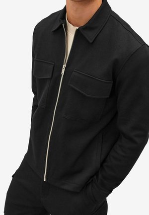 Mand iført en sort zip-up jakke med to klaplommer på brystet og krave over en hvid skjorte, hænderne i lommerne.