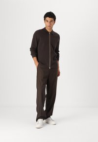 Pulover crem cu fermoar, cu guler și buzunar frontal, asociat cu pantaloni maro lejeri și adidași albi. Design simplu, modern.