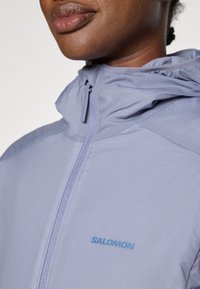 Lys lilla hettegenser laget av mykt stoff, med frontglidelås og en blå "SALOMON" logo på venstre bryst.