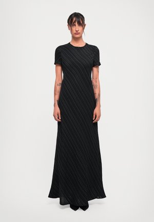 Mujer con tatuajes que lleva un vestido negro de manga corta y largo hasta el suelo, con finas rayas diagonales blancas, de pie contra un fondo claro y sencillo.