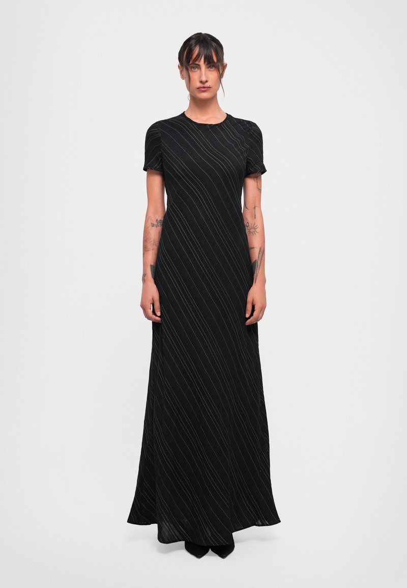 Woman with tattoos wearing a black, short-sleeve, floor-length dress with thin white diagonal stripes, standing against a plain light background.