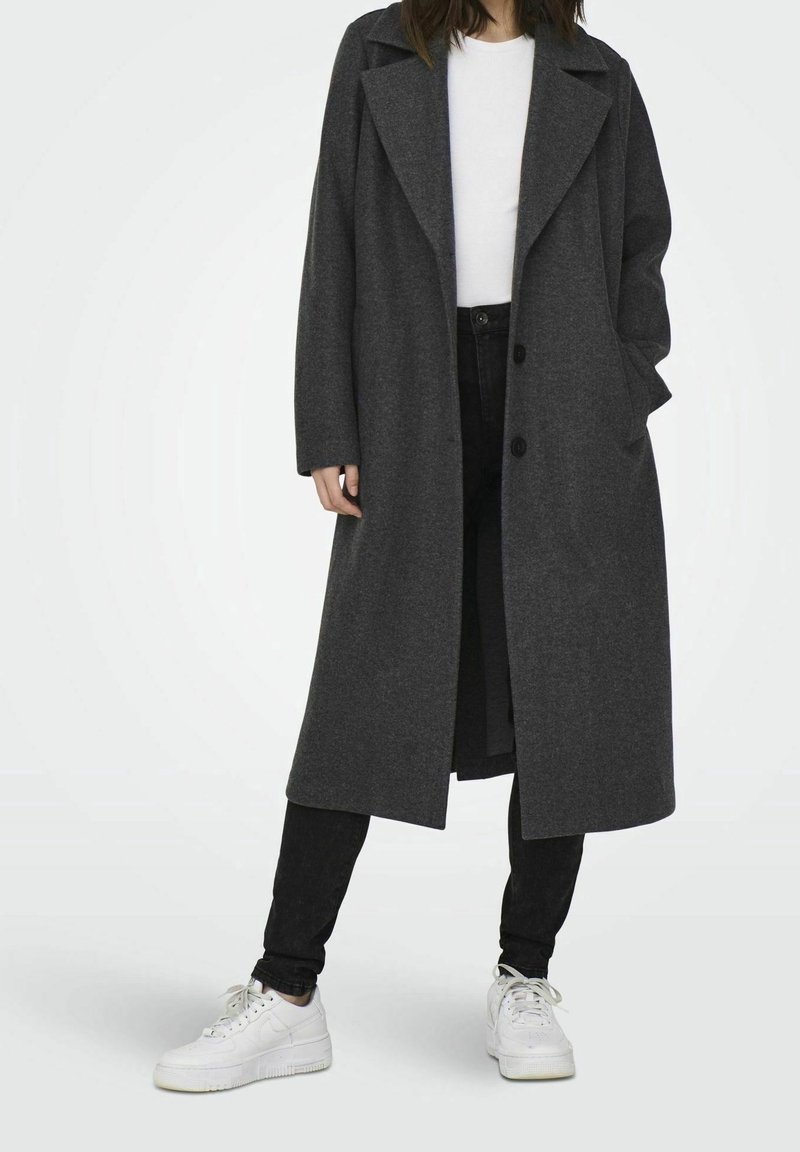 Grijze wolblend lange jas met een notch-kraag, dubbele knoopsluiting, zijzakken en een relaxte pasvorm, gecombineerd met zwarte jeans en witte sneakers.