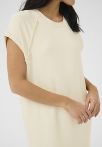 Rochie scurtă, de culoare bej deschis, cu mâneci scurte și o croială lejeră, realizată dintr-un material moale. Prezintă un guler rotund și detalii subtile de cusătură.