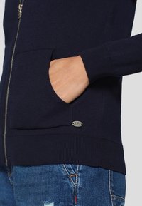 Marineblauwe ritstrui met een gladde textuur, ribbelmanchetten en -zoom. Voorzien van een voorzak en een metalen logo-accent nabij de zoom.