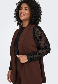 Brown sleeveless vest with a flowing design, worn over a black long-sleeve blouse featuring floral lace patterns. Smooth fabric and relaxed fit.