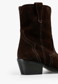 Bruine suède enkellaars met een kleine hak, voorzien van contrasterende stiksels en zijuitsparingen. Het ontwerp benadrukt een door het Westen geïnspireerde stijl.