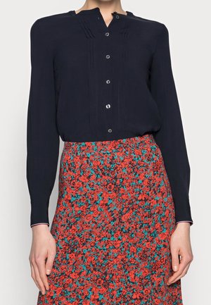 Woman wearing a long-sleeve black buttoned blouse tucked into a red floral patterned skirt. Hands relaxed by sides.