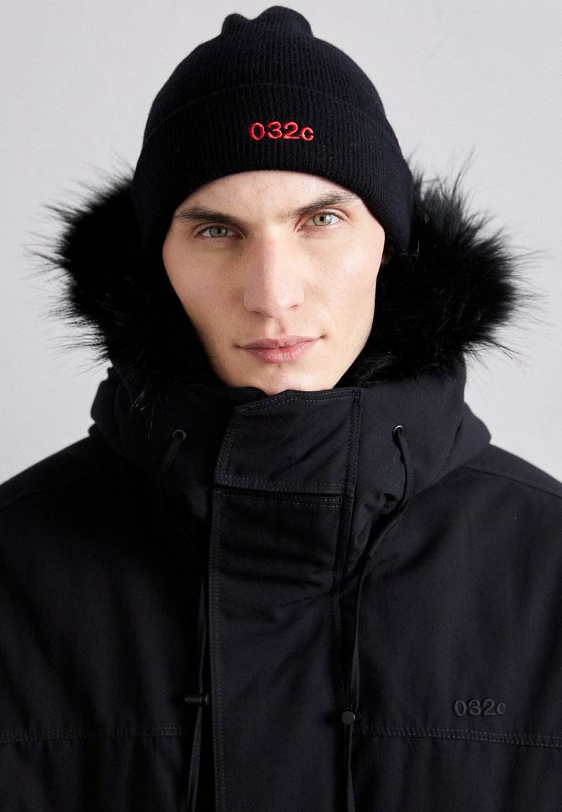 Schwarze Pufferjacke mit hohem Kragen, Fellbesatz und gesticktem "032c"-Logo; kombiniert mit einer schwarzen gerippten Mütze. Strukturiertes Material sichtbar.