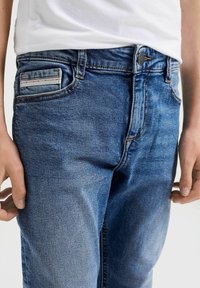 Los jeans de denim azul desgastado presentan un diseño clásico de cinco bolsillos, con un sutil acento de rayas rojas en el bolsillo trasero y un cierre de botón.