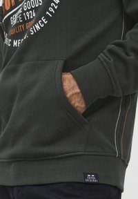 Sudadera de color verde oscuro hecha de una mezcla de algodón, con un bolsillo frontal, puños acanalados y un logo bordado en blanco y naranja.