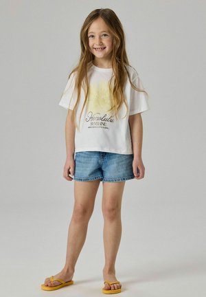 Smilende pige med langt hår iført hvid T-shirt med gul blomsterprint, denimshorts og gule flip-flops, der står mod en ensfarvet baggrund.