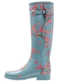 Bota de goma brillante y azul hasta la rodilla, que presenta un patrón floral con flores rosas y ramas marrones. Detalle de hebilla en el lateral.
