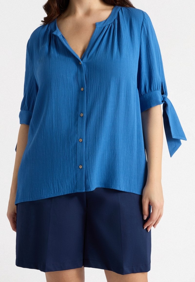Blouse bleue boutonnée avec une finition texturée, manches retroussées nouées aux poignets et encolure en V. Associée à un short bleu marine.