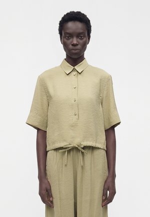 Femme portant une chemise ample beige à manches courtes boutonnée avec col et un pantalon taille haute assorti avec cordon, sur fond blanc.