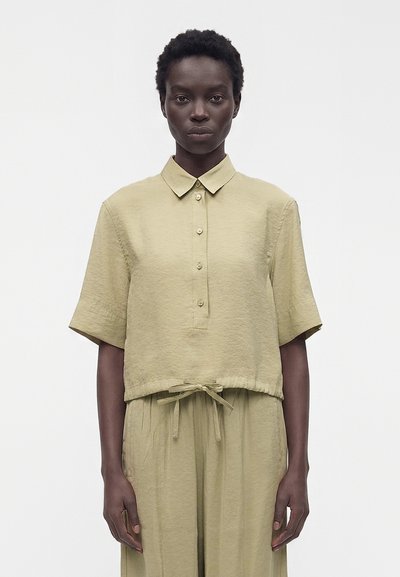 Femme portant une chemise ample beige à manches courtes boutonnée avec col et un pantalon taille haute assorti avec cordon, sur fond blanc.