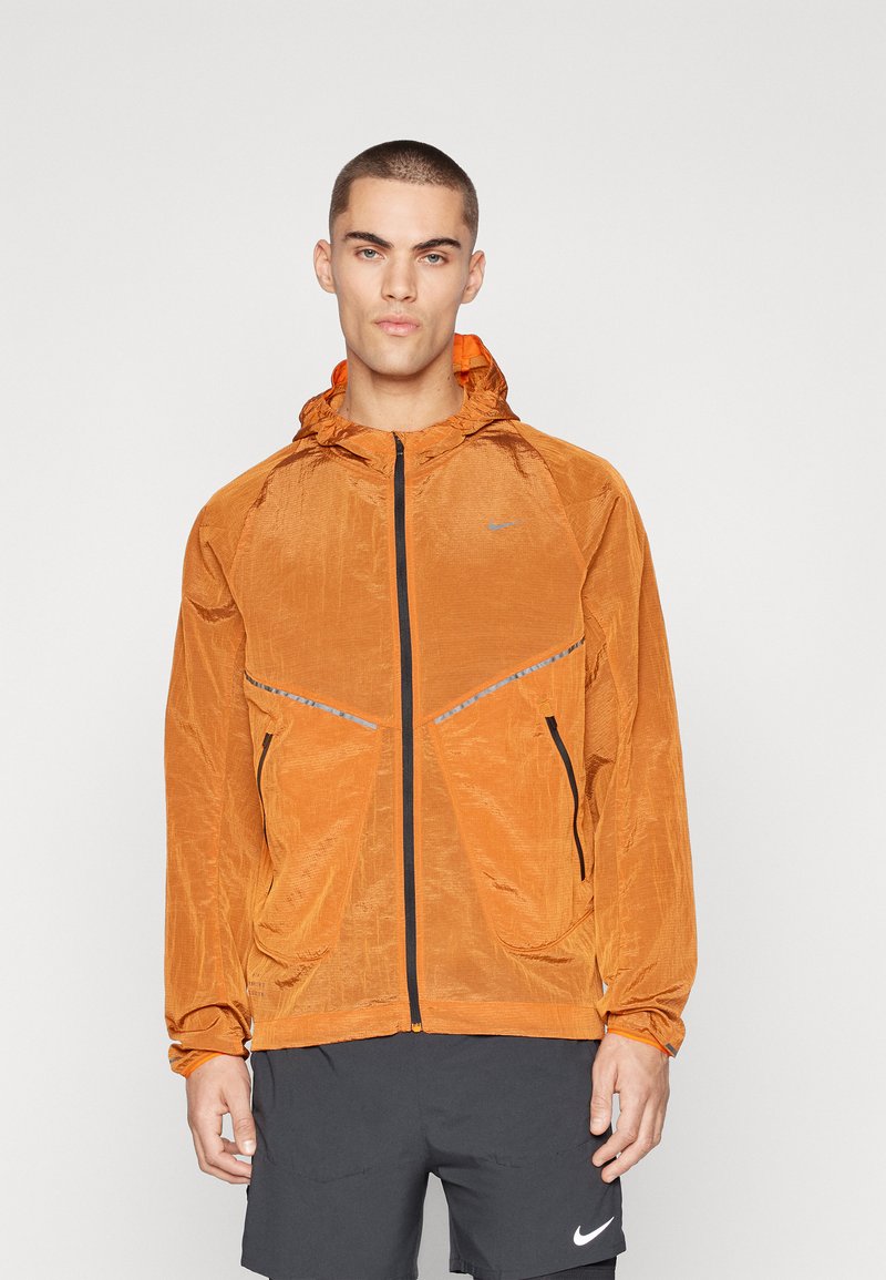 Oranje lichtgewicht jack met een textuurafwerking, een ritssluiting aan de voorkant en een capuchon. Bevat reflecterende accenten en zwarte ritszakken.