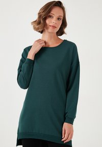 Puhafabric-ból készült, türkiz színű, hosszú ujjú felső, kerek nyakkivágással és bordás mandzsettákkal, laza szabással és oldalsó hasítékokkal az alján.