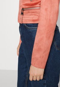 Korálová bunda z umělé semiše s kapsou na zip a vodorovným prošíváním. Nositelná přes tmavě modré džínové kalhoty. Hladký povrch a přiléhavý střih.