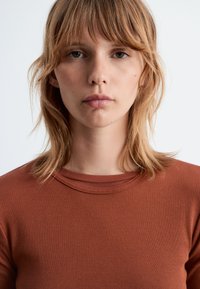 Brauner gerippter Langarm-Top mit Rundhalsausschnitt, der eine glatte Textur und eine figurbetonte Passform aufweist, was einen minimalistischen Stil betont.