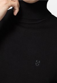 Pull à col roulé noir en matière côtelée, avec un logo subtil au centre. Vue en gros plan mettant en valeur la texture et la coupe.