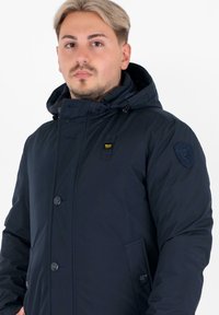 Giacca puffer blu navy con cappuccio, chiusura con bottoni e tasche laterali. Presenta un badge cucito sul braccio sinistro e una texture liscia.