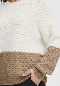 Strickpullover mit einem weißen oberen Abschnitt und einem strukturierten braunen unteren Teil, mit gerippten Bündchen und Saum. Hand vor der Tasche platziert.