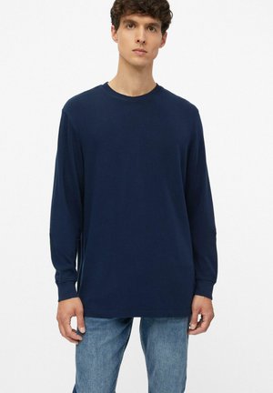 Junger Mann, der ein schlichtes dunkelblaues Langarmshirt und hellblaue Jeans trägt, steht vor einem weißen Hintergrund.