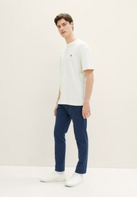Vit kortärmad T-shirt med en liten svart logotyp, kombinerad med mörkblå byxor och vita sneakers. Modellen står mot en neutral bakgrund.