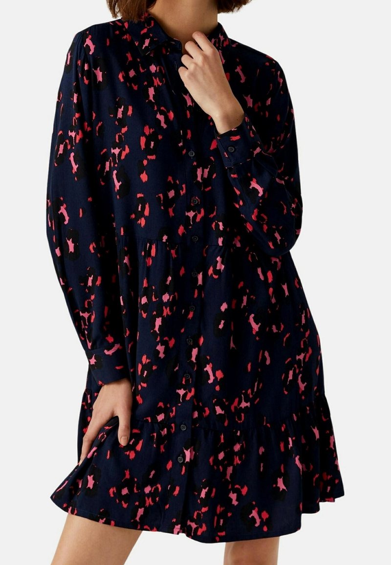 Femme portant une robe chemise ample bleu foncé à manches longues avec un motif abstrait rouge et rose, tenant l'ourlet de la robe d'une main.