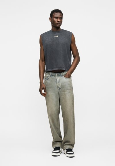 Άνδρας μοντέλο φορεί ένα αμάνικο φθαρμένο μαύρο τοπ, φαρδιά φθαρμένα γκρι τζιν και μαύρα-άσπρα αθλητικά παπούτσια, στέκεται με το ένα χέρι στην τσέπη.