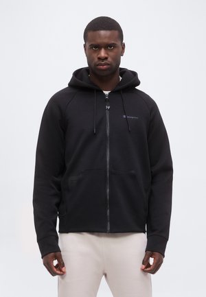 Mann trägt einen schwarzen Kapuzenpullover mit Reißverschluss und heruntergelassener Kapuze sowie beige Hose, steht frontal vor einem einfarbigen hellen Hintergrund.
