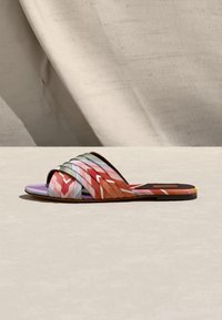 Sandale plate à slide avec des brides tissées multicolores aux motifs rouge, orange, rose, violet et vert sur une surface neutre.