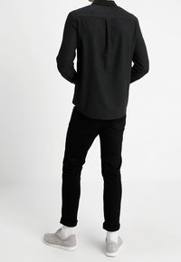 Zwart shirt met lange mouwen en een knoopsluiting bij de kraag, met een plooi aan de achterkant, gecombineerd met zwarte jeans en grijze sneakers met witte zolen.