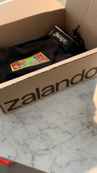 Prenda de ropa negra con un parche colorido que presenta un diseño de dragón, dentro de una caja de cartón de Zalando sobre una superficie de mármol.