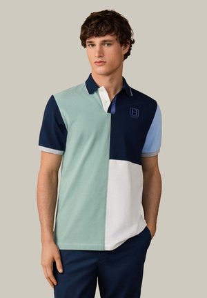 Joven con una camisa polo de bloques de colores en azul marino, azul claro, verde menta y blanco, de pie con una mano en el bolsillo.