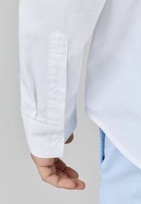 Poignet de chemise blanche à boutons, avec une conception structurée et un tissu intérieur bleu clair contrastant au poignet.